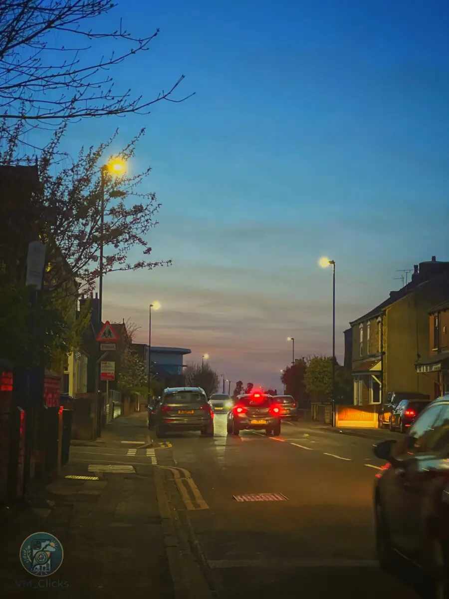 Dusk street scene captured on iPhone 13 Pro in Mexborough, UK — warm streetlights glow against a blue evening sky with car brake lights on a busy road, colour graded in Snapseed